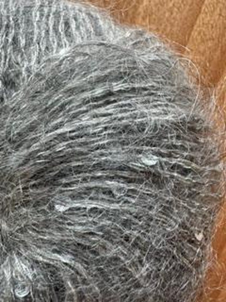 May include: Close-up of a ball of gray yarn with a textured, slightly fuzzy appearance. The yarn appears to be a blend of gray and white fibers, creating a heathered effect. The fibers are tightly wound, and the overall texture is soft and inviting.