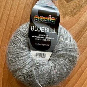 May include: A ball of Sesia Bluebell yarn, primarily gray with subtle silver highlights, sits on a wooden surface. The yarn label displays the brand name, yarn type, and fiber content: 42% Mohair, 34% Polyester, 24% Silk.