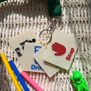 May include: A green lanyard with a sunflower pattern attached to a metal ring holding several square white communication cards. The cards feature raised icons and text including a red hand with the word Stop and blue text reading Drink.