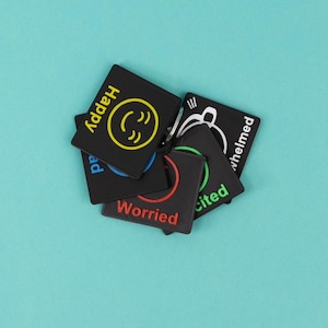 May include: A collection of square black keychains featuring colourful text and icons representing emotions. The visible labels include Happy in yellow, Worried in red, Excited in green, and Overwhelmed in white, each paired with a matching graphic.