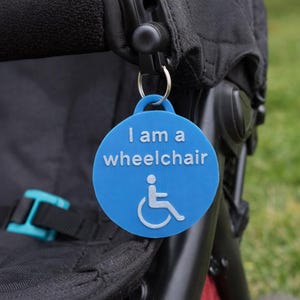 May include: A circular blue plastic tag attached to a black pushchair frame by a metal ring. The tag features the white embossed text "I am a wheelchair" above a white wheelchair accessibility symbol, clearly identifying the pushchair as a wheelchair.