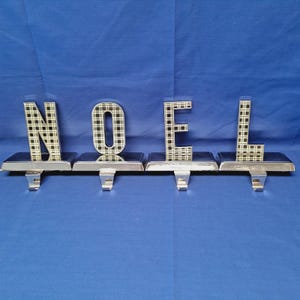 May include: Silver-toned metal stocking holders spelling "NOEL". Each letter is a different stocking holder. The letters are filled with a black and white plaid pattern. The holders have a hook for hanging stockings.