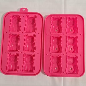 Op de afbeelding: Twee felroze siliconen mallen, elk met zes katvormige holtes. De mallen zijn ontworpen voor het maken van traktaties of knutselwerkjes. Elke kattenmal heeft een gedetailleerd gezicht en lichaam. De mallen liggen op een wit oppervlak.