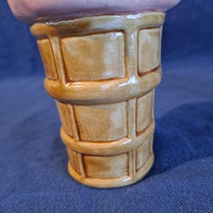 K&ouml;nnte beinhalten: Ein keramischer Eisbecher in Form eines Eiswaffelkegels mit einem hellbraunen Waffelmuster. Der obere Teil ist rosa-lila gef&auml;rbt und &auml;hnelt Eiscreme. Der Beh&auml;lter ist gl&auml;nzend und scheint ein dekorativer Gegenstand zu sein.