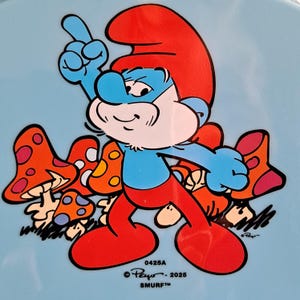 May include: A Smurf cartoon character with a red hat and pants, blue skin, and a white beard, pointing upwards. The character is surrounded by orange and white mushrooms on a light blue background. The text "0425A", "&copy; Peyo - 2025", and "SMURF&trade;" are visible.