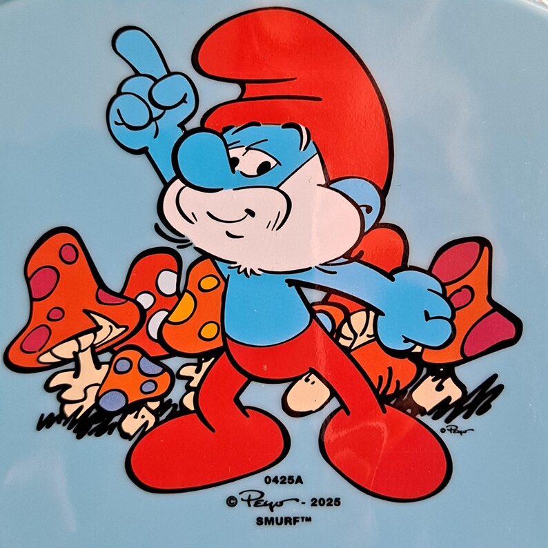 May include: A Smurf cartoon character with a red hat and pants, blue skin, and a white beard, pointing upwards. The character is surrounded by orange and white mushrooms on a light blue background. The text "0425A", "&copy; Peyo - 2025", and "SMURF&trade;" are visible.