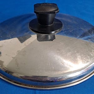 May include: A stainless steel pot lid with a black handle. The lid is round with a brushed metal finish and a black plastic handle. The handle is square-shaped and has the word "non-stick" printed on it.