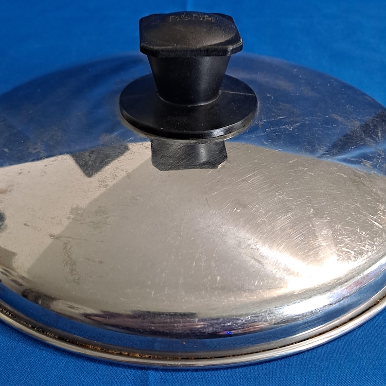 May include: A stainless steel pot lid with a black handle. The lid is round with a brushed metal finish and a black plastic handle. The handle is square-shaped and has the word "non-stick" printed on it.