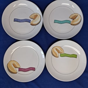 Puede incluir: Cuatro platos de cerámica blanca con borde negro y una galleta de la fortuna en cada uno. Las galletas de la fortuna tienen diferentes refranes: "¿Por qué no asumir la responsabilidad de tu grandeza?", "Lo que das, recibes lo que quieres", "Piensas que es un secreto, pero no lo es", y "El amor vale la pena."