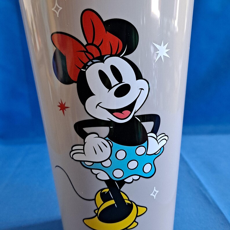 May include: A tall, light grey tumbler with a smiling Minnie Mouse illustration. Minnie is in her classic outfit: a red bow, black top, blue polka-dot skirt, and yellow shoes. The tumbler has star and square accents.