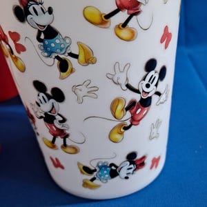 Puede incluir: Vaso blanco con im&aacute;genes de dibujos animados de Mickey y Minnie Mouse. El vaso tiene un patr&oacute;n repetido de los personajes en varias poses, con lazos y guantes rojos. El fondo es azul liso.