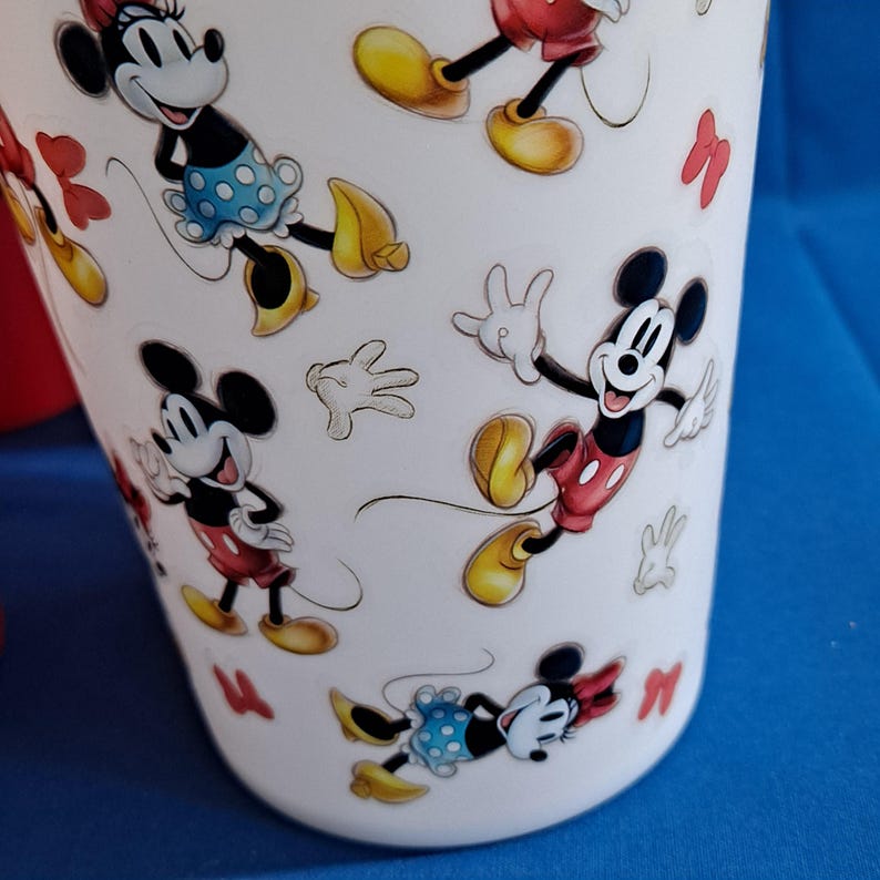 Puede incluir: Vaso blanco con im&aacute;genes de dibujos animados de Mickey y Minnie Mouse. El vaso tiene un patr&oacute;n repetido de los personajes en varias poses, con lazos y guantes rojos. El fondo es azul liso.