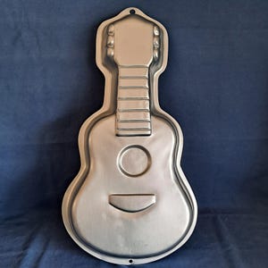 Puede incluir: Un molde para pastel de metal plateado con forma de guitarra. El molde tiene un diseño detallado de una guitarra, incluyendo las cuerdas, el agujero de sonido y la golpeadora.