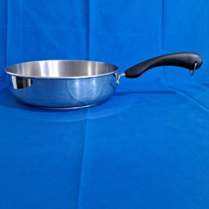 Puede incluir: Una sartén de acero inoxidable con un mango de goma negro. La sartén tiene una superficie brillante y reflectante.