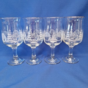 May include: Four clear wine glasses with a winter scene etched on the front. The design features snow-covered trees and falling snowflakes. Each glass has a gold rim and a stem.