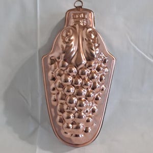 May include: Copper-colored metal mold in the shape of a bunch of grapes. The mold has a decorative design with a loop at the top for hanging. The grapes are detailed with individual spheres, and the leaves are also detailed.
