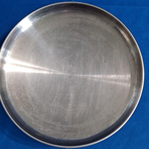 May include: A round, stainless steel plate with a brushed metal finish. The plate has a slightly raised edge and a smooth, reflective surface. The plate is designed for serving food.