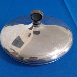 Pode incluir: Uma tampa de metal prateado brilhante com um botão preto na parte superior. A tampa é redonda com uma superfície ligeiramente curva. O botão é uma pequena esfera escura. A tampa parece ser para uma panela ou frigideira.