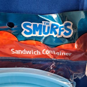 May include: A blue and orange plastic sandwich container featuring the text "THE SMURFS" in white and blue. The words "Sandwich Container" are printed below the logo.
