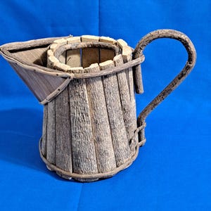 May include: A rustic pitcher crafted from weathered wood, featuring a curved handle and a spout. The body is constructed from vertical wooden planks, bound together with a thin, woven material. The pitcher has a natural, earthy aesthetic.