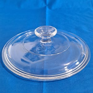 May include: A clear glass lid with a round knob handle. The lid is circular with a slightly raised center and a rim. The knob is also clear and round. The lid is likely for a pot or pan.