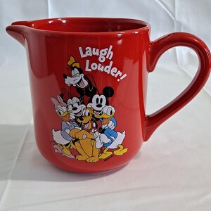 May include: A red ceramic pitcher featuring Disney characters, including Mickey Mouse, Minnie Mouse, Donald Duck, and Goofy. The pitcher has a large handle and a spout. The words "Laugh Louder!" are printed on the side.