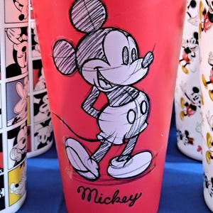 Puede incluir: Vaso rojo con un boceto de Mickey Mouse y la palabra "Mickey" en cursiva. Otros vasos con dise&ntilde;os de Mickey Mouse son visibles. El vaso es cil&iacute;ndrico y parece ser de pl&aacute;stico.
