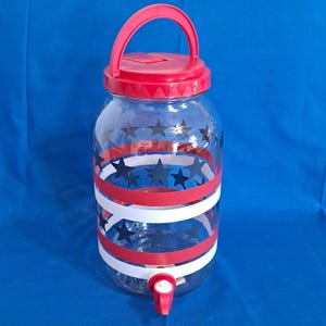 May include: A clear glass beverage dispenser with a red lid, handle, and spigot. The jar is decorated with red, white, and black star and stripe designs. The dispenser is designed for cold beverages.