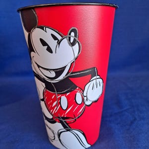 Puede incluir: Un vaso alto rojo con un borde negro, con un gr&aacute;fico de Mickey Mouse sonriente. El vaso tiene un fondo rojo vibrante. Mickey Mouse est&aacute; representado en su esquema de color cl&aacute;sico negro, blanco y rojo, con una expresi&oacute;n juguetona.