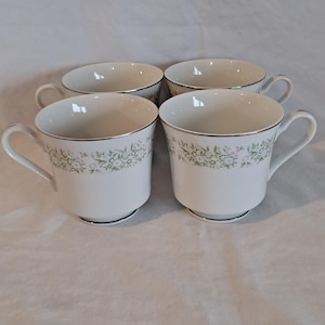 Puede incluir: Juego de cuatro tazas de té de cerámica blanca con un diseño floral y un borde plateado. Las tazas tienen un delicado patrón floral en tonos verdes, rosas y blancos.