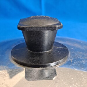 May include: Close-up of a stainless steel pot lid with a black plastic handle. The handle is square-shaped at the top and tapers down to a circular base. The lid has a shiny, reflective surface.