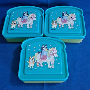 Puede incluir: Tres fiambreras turquesas con forma de sándwich. Cada tapa presenta una imagen de dibujos animados de un perro azul montando a caballo, con un personaje de perro más pequeño. La palabra "zak!" está impresa en cada tapa.