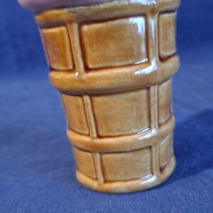 K&ouml;nnte beinhalten: Ein keramischer Beh&auml;lter in Form einer Eist&uuml;te. Die Basis ist ein hellbrauner Waffelkegel, und die Oberseite ist rosa-lila. Der Kegel hat eine gl&auml;nzende Oberfl&auml;che und ein detailliertes, strukturiertes Design.