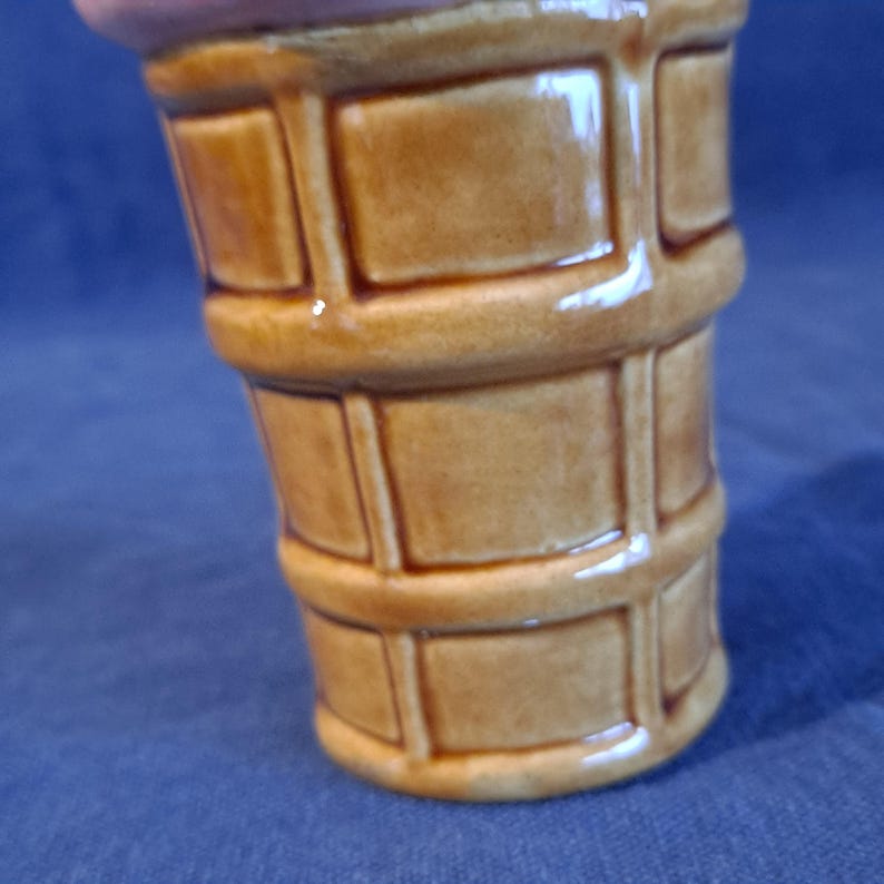 K&ouml;nnte beinhalten: Ein keramischer Beh&auml;lter in Form einer Eist&uuml;te. Die Basis ist ein hellbrauner Waffelkegel, und die Oberseite ist rosa-lila. Der Kegel hat eine gl&auml;nzende Oberfl&auml;che und ein detailliertes, strukturiertes Design.