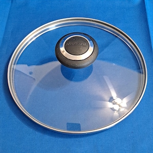 May include: A clear glass Farberware lid with a stainless steel rim and a black and silver handle. The lid is circular and sits on a blue surface. The Farberware logo is visible on the handle.