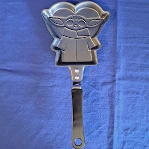 Może przedstawiać: Srebrna metalowa forma do naleśników w kształcie postaci z popularnej serii science fiction. Forma ma długi czarny uchwyt z otworem na końcu do zawieszania. Twarz i ubranie postaci są szczegółowe.