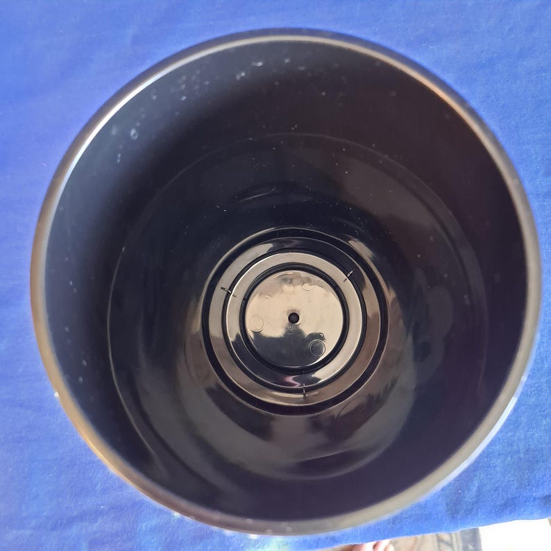 Puede incluir: Una maceta de pl&aacute;stico negro, vista desde arriba, que muestra los detalles interiores. La maceta tiene un dise&ntilde;o circular con anillos conc&eacute;ntricos en la parte inferior. La maceta est&aacute; sobre una superficie azul.