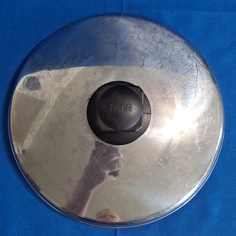 May include: A stainless steel pot lid with a black plastic handle. The lid is circular with a shiny, reflective surface. The handle is centered and has the word "Rona" embossed on it.