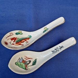 Könnte beinhalten: Zwei asiatische Suppenlöffel aus weißer Keramik mit goldenem Rand. Einer der Löffel zeigt ein rotes und grünes Drachendesign, der andere eine Person in einer Landschaft. Die Löffel liegen auf einer blauen Oberfläche.