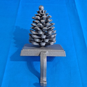 May include: A gray metal stocking holder shaped like a pine cone. The pine cone sits atop a rectangular base with a hook for hanging. The holder is made of cast iron and is designed to hold a Christmas stocking. The background is a solid blue.