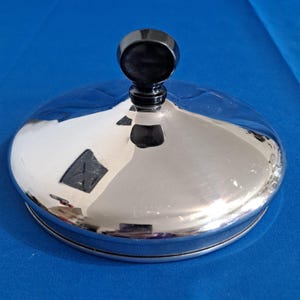 Pode incluir: Uma rolha de garrafa prateada brilhante com um topo preto arredondado. A superfície da rolha é reflexiva, refletindo o fundo azul. A rolha é redonda e parece ser feita de metal.