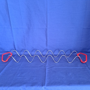 May include: A silver-colored taco holder with a wavy design. The holder has two red handles on each end. The taco holder is designed to hold multiple tacos upright, preventing them from falling over. The background is a solid blue color.