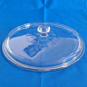 May include: A clear, oval-shaped glass lid with a square knob. The lid is transparent, allowing a view of what's beneath. The knob is also clear, and the lid has a slightly raised edge. The background is a solid blue.