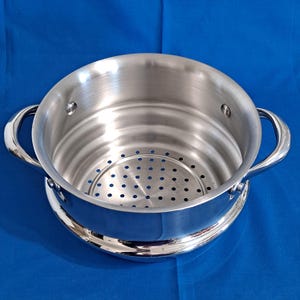 May include: A stainless steel steamer pot with a perforated insert and two curved handles. The pot sits on a slightly raised base and is set against a blue background. The interior of the pot is visible.