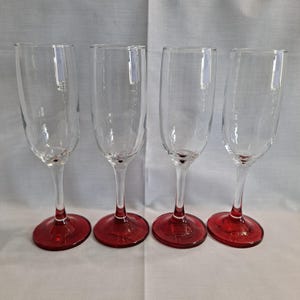 May include: Four clear champagne flutes with red bases. The glasses have a tall, slender bowl and a long stem. The red base is circular and adds a pop of color to the elegant design. Perfect for celebrations.