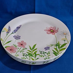 Puede incluir: Un juego de cuatro platos de cena de cerámica blanca con un diseño floral. Los platos presentan una variedad de flores rosas, moradas y amarillas con hojas verdes.