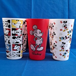 Puede incluir: Un conjunto de vasos altos y cil&iacute;ndricos con dise&ntilde;os de Mickey Mouse. Un vaso rojo tiene una gran ilustraci&oacute;n de Mickey Mouse con el nombre "Mickey" debajo. Otros vasos tienen patrones repetidos de Mickey Mouse en varias poses y colores.