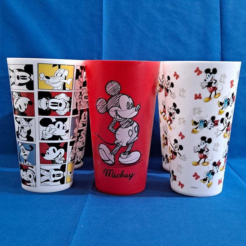 Puede incluir: Un conjunto de vasos altos y cil&iacute;ndricos con dise&ntilde;os de Mickey Mouse. Un vaso rojo tiene una gran ilustraci&oacute;n de Mickey Mouse con el nombre "Mickey" debajo. Otros vasos tienen patrones repetidos de Mickey Mouse en varias poses y colores.