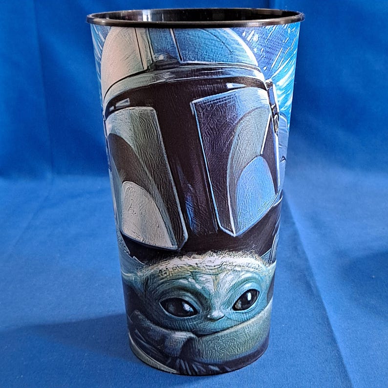 May include: A tall, plastic tumbler featuring a detailed illustration of a Mandalorian helmet and Grogu, also known as Baby Yoda, in shades of blue, black, and gray. The cup has a black rim and a blue background.