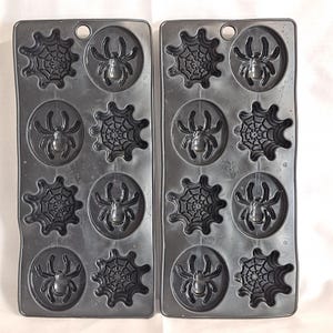 May include: Two dark grey plastic Halloween-themed moulds. Each mould has six cavities with spider and spiderweb designs. The moulds are designed for making chocolates, sweets, or other treats for the holiday.